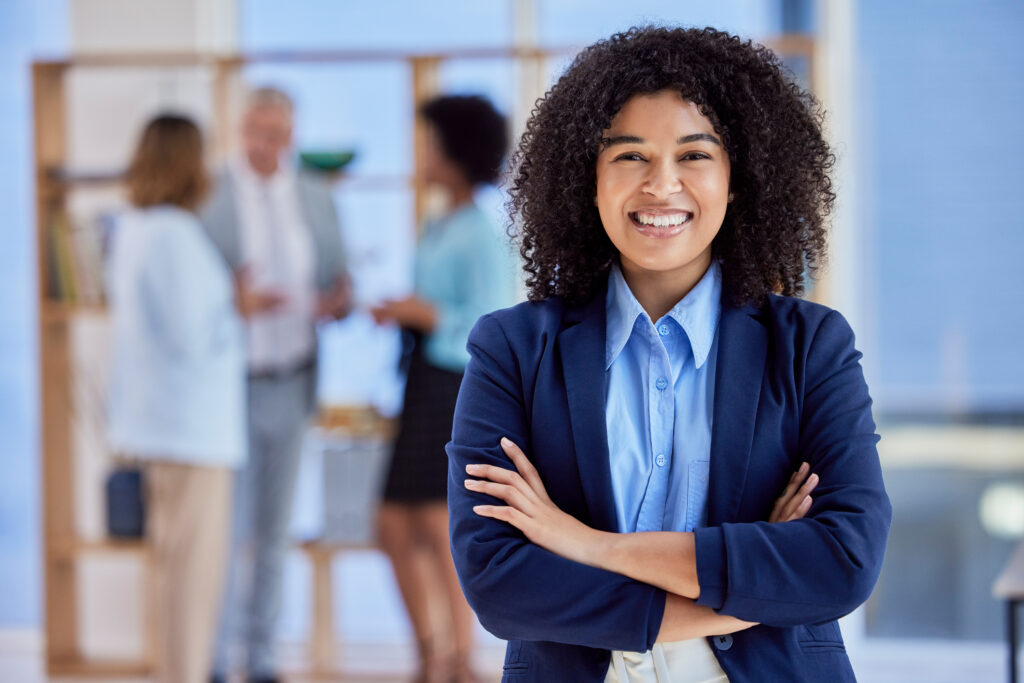 A smiling young professional standing with her arms crossed while her colleagues in the background